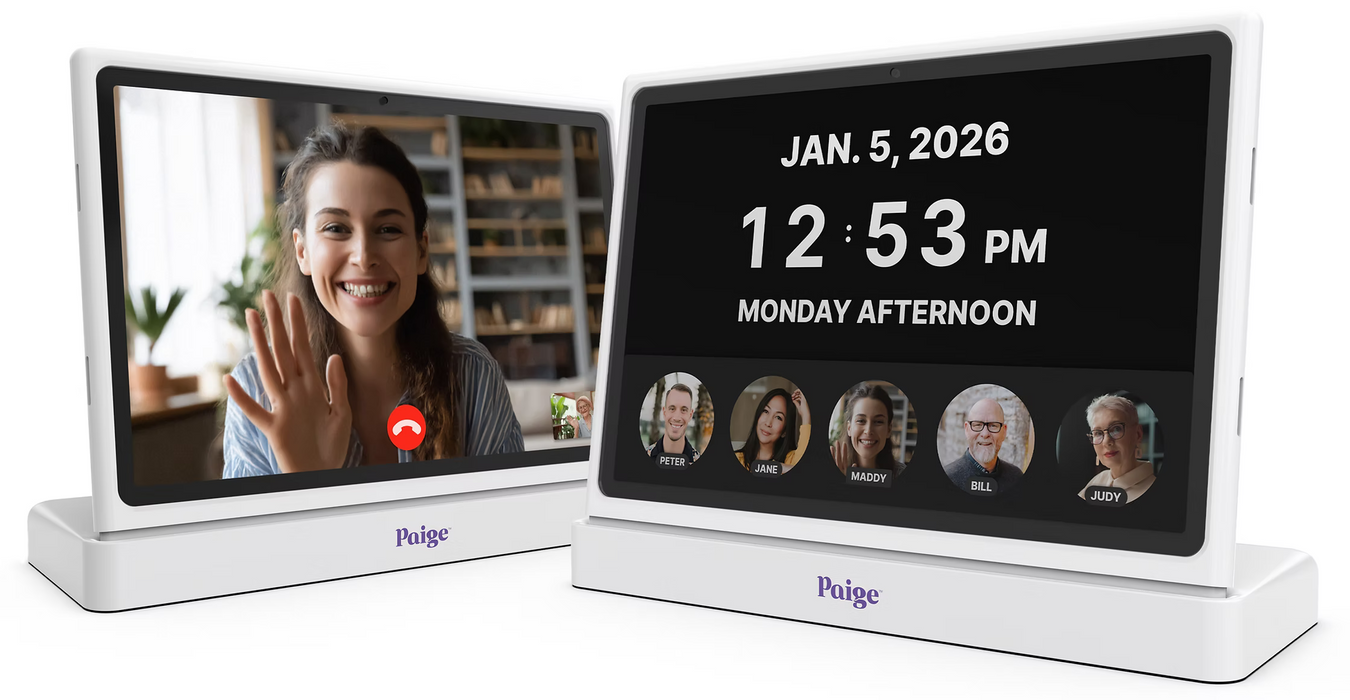 Two white digital video display screens on matching white bases. The front screen displays the date and time, and the side screen shows a video call interface with a woman waving and the Paige logo at the bottom.