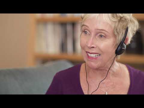 A video in which a grey-haired lady sits talking with a child as they play with a toy.  She is using a SuperEar personal sound amplifier to hear them speak. 