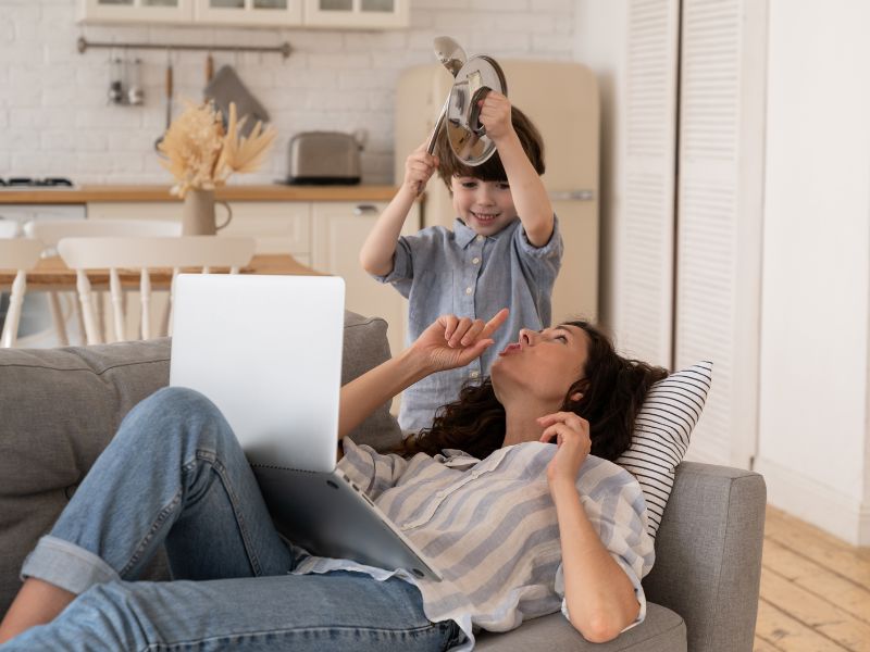 Woman lying on a couch with a laptop, child playfully interacting above her.
