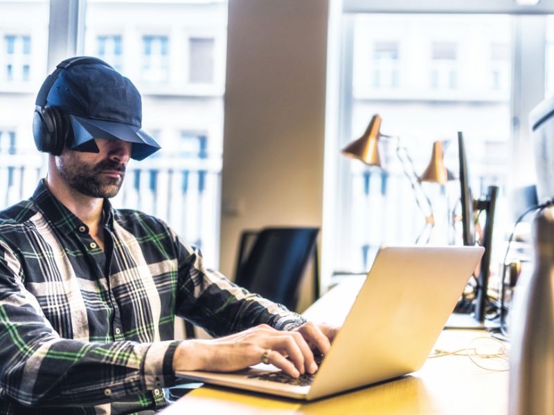 Person wearing a hat with side blinders and headphones, working on a laptop in an office setting.