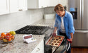 Phot of a woman removing burnt cookie from the oven and the main receiver screen displays the word SMOKE.  Sonic Alert HomeAware Smoke & CO Sound Signaler HA360SSSCK2-1 